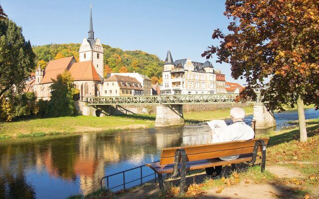 Älterer Mann auf Bank am Fluss, liest Zeitung; im Hintergrund Brücke und Kirche.