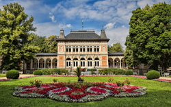 Historisches Gebäude mit Erker, umgeben von Bäumen und Blumenbeeten im Vordergrund.