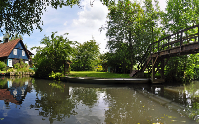 Fachwerkhaus an einem Teich mit Holzbrücke und üppiger Vegetation.