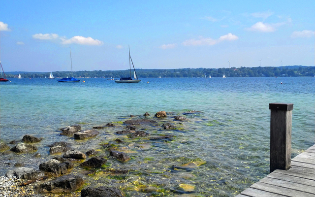 Steg am klaren See mit Segelbooten am Horizont unter blauem Himmel.