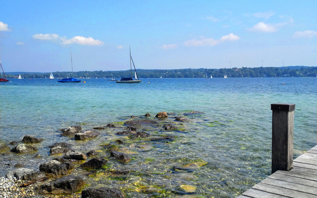 Steg am klaren See mit Segelbooten am Horizont unter blauem Himmel.