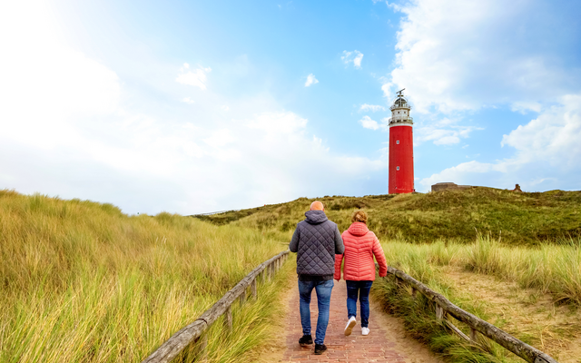 Zwei Personen gehen einen Weg entlang zu einem roten Leuchtturm in Dünenlandschaft.
