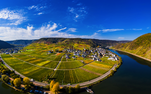 Panoramablick auf eine Flusslandschaft mit einem Dorf und Weinbergen unter blauem Himmel.