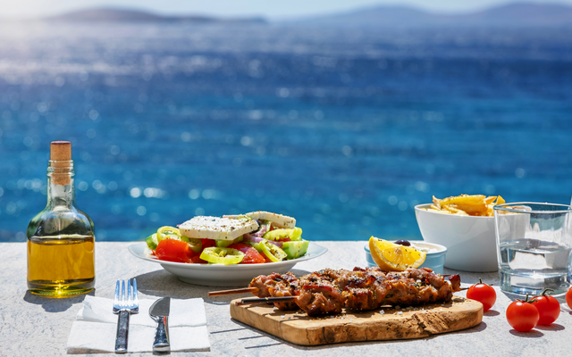 Tisch mit Grillspießen, Salat und Getränken vor Meereshintergrund.