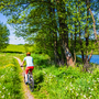 Radfahrerin auf einem Radweg entlang eines Sees und zwischen grünen Wiesen im Landschaftspark Suwalski, Polen