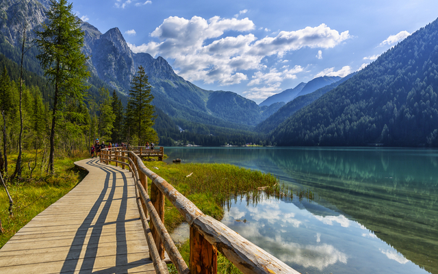 Holzsteg führt entlang eines klaren Bergsees mit Bergen und Bäumen im Hintergrund.