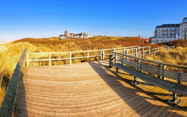 Holzsteg durch Sanddünen mit Meer und Hotel im Hintergrund bei klarem Himmel