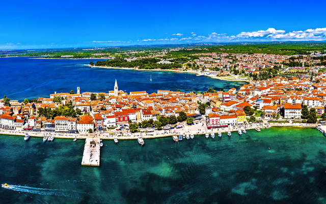 Küstenstadt mit roten Dächern und Hafen vor blauem Meer und Himmel.