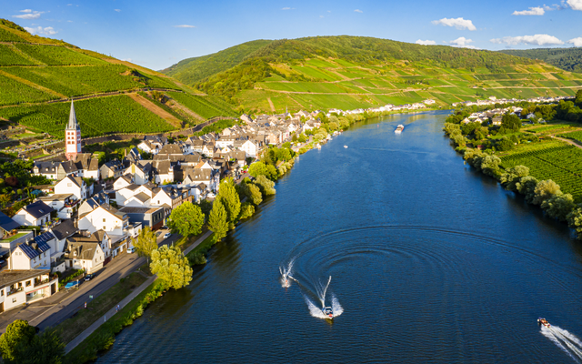 Dorf am Fluss mit Weinbergen im Hintergrund und Booten auf dem Wasser.
