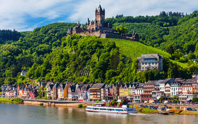 Burg auf Hügel über kleiner Stadt am Flussufer, mit bewaldetem Hintergrund.