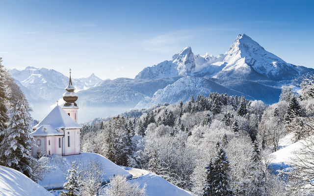 Verschneite Kirche vor malerischen Alpenpanorama bei klarem Himmel.