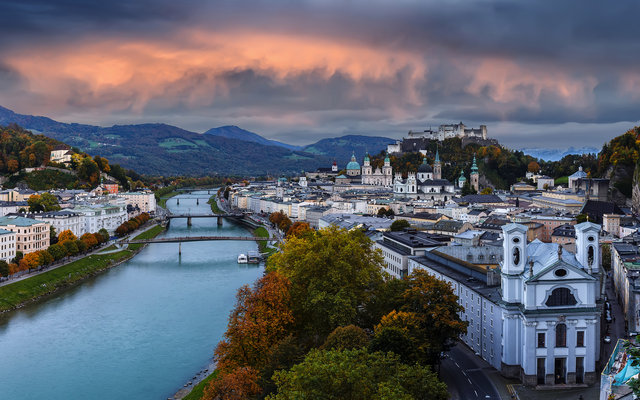 Panoramablick auf eine Stadt mit Fluss, Brücke, historischen Gebäuden und bewölktem Himmel.