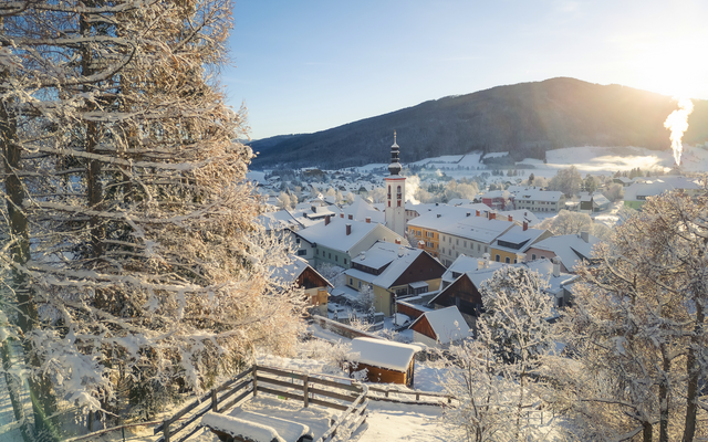 Winterlandschaft mit verschneitem Dorf und Kirchturm im Sonnenlicht.