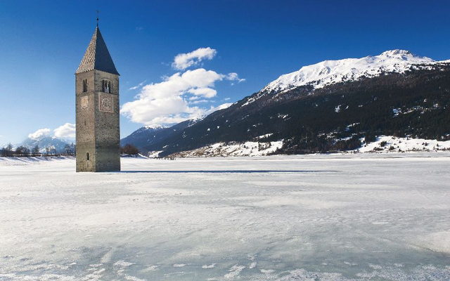 Kirchturm im zugefrorenen Reschensee mit schneebedeckten Bergen im Hintergrund.