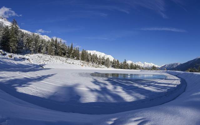 Verschneite Berglandschaft mit zugefrorenem See und Nadelbäumen unter klarem Himmel.