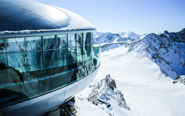 Modernes Gebäude auf schneebedecktem Berggipfel mit Panorama-Ausblick auf Alpen.