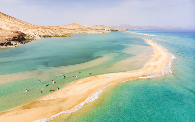 Luftaufnahme eines Strandes mit türkisblauem Wasser und Windsurfern.