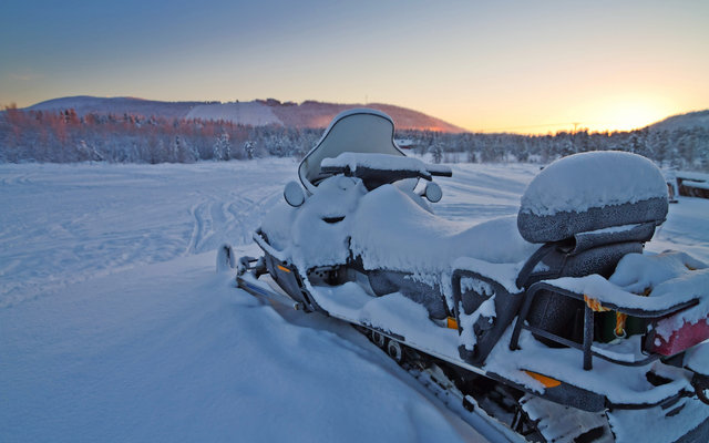 Ein schneebedecktes Schneemobil in winterlicher Landschaft bei Sonnenuntergang.