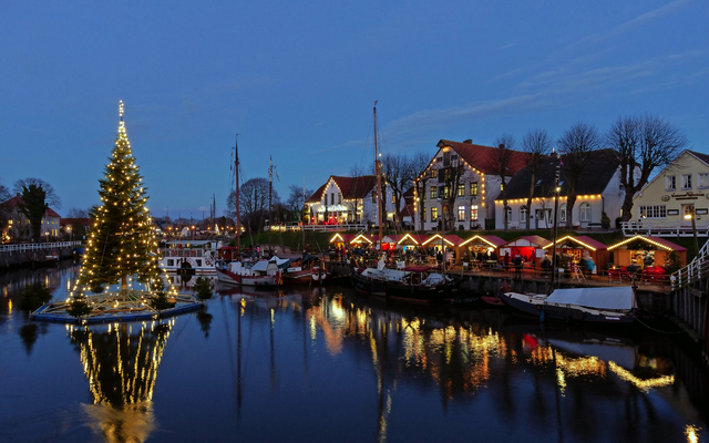 Weihnachtsbaum beleuchtet Hafen bei Nacht, Boote und Gebäude im Hintergrund.