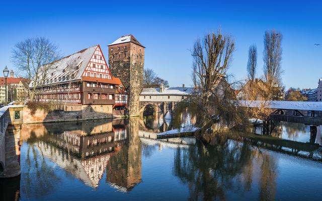 Fachwerkhaus und Brücke an verschneitem Flussufer, klarer Himmel, spiegelndes Wasser.