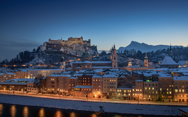 Verschneite Stadt und Burg bei Abenddämmerung, beleuchtet entlang eines Flusses.