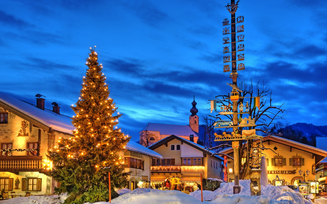Beleuchteter Weihnachtsbaum auf verschneitem Platz mit Kirche und Häusern.