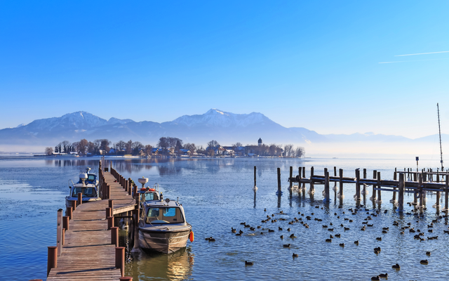 Hölzerner Steg mit Booten auf einem See, Berge im Hintergrund, viele Wasservögel.
