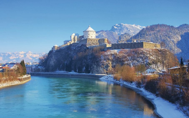 Burg auf einem Hügel an einem Fluss im winterlichen Alpenpanorama.