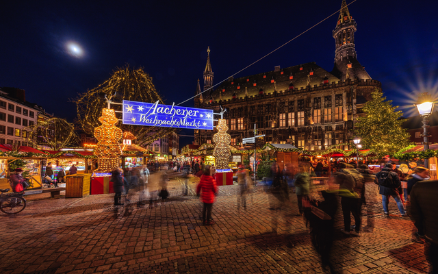 Nachtaufnahme eines belebten Weihnachtsmarktes mit dekorierten Ständen und Lichtern.