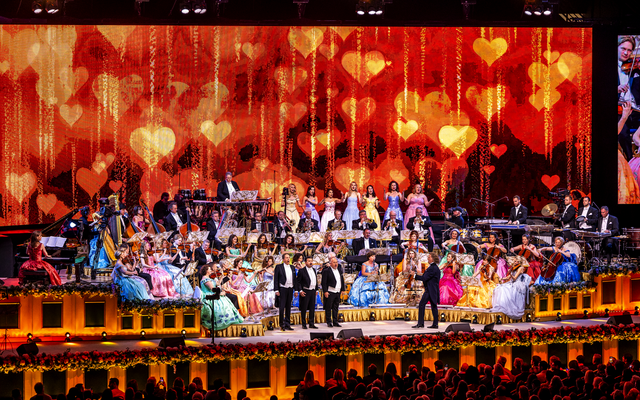 Orchester spielt vor Bühne mit Herzdekor, viele Künstler und bunte Kleider.