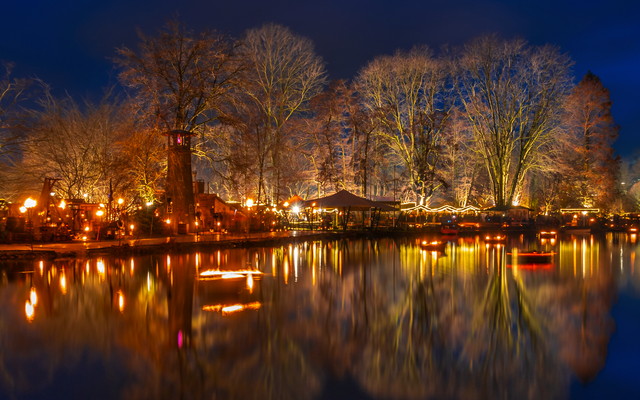 Ein stimmungsvoll beleuchteter Park am Abend mit Bäumen und deren Spiegelbild in einem ruhigen See.