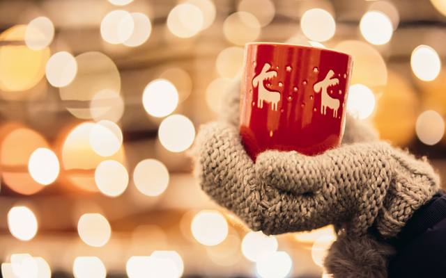 Hände in Handschuhen halten Tasse mit Rentiermotiv vor Lichterhintergrund.