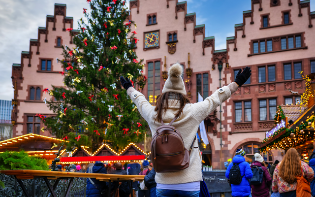 Frau vor Weihnachtsmarkt und geschmücktem Baum in Stadt mit historischen Gebäuden.