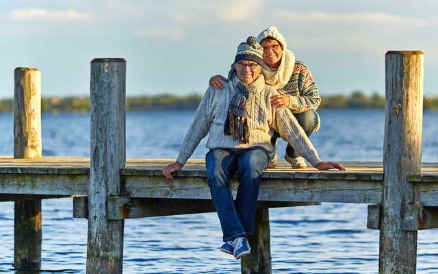 Zwei Menschen in Winterkleidung sitzen auf einem Steg über einem See.