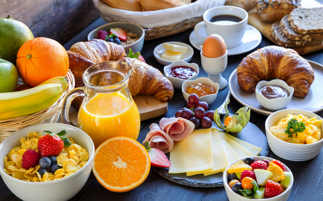 Reichhaltiges Frühstück mit Saft, Croissants, Obst, Müsli und Käse auf einem Tisch.