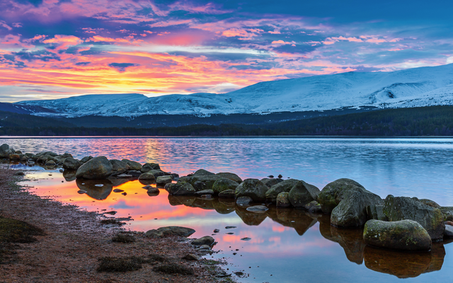 Felsige Küste an einem See bei Sonnenuntergang mit schneebedeckten Bergen im Hintergrund.