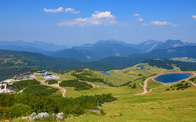 Panorama von Alpenlandschaft mit Hügeln, Wäldern und kleinem See unter blauem Himmel.
