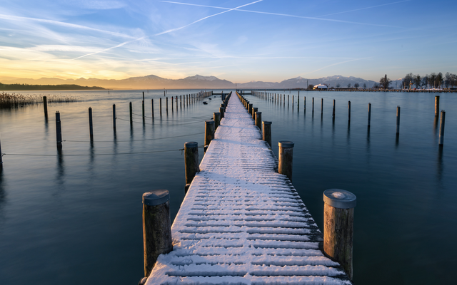 Schneebedeckter Steg führt bei Sonnenuntergang über stillen See.