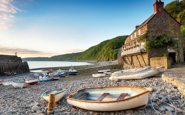 Boote liegen an einem steinigen Strand neben einem alten Haus, mit bewaldeten Hügeln im Hintergrund.