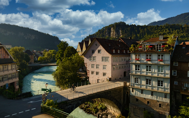 Stadtansicht mit Brücke über Fluss, umgeben von historischen Gebäuden und grüner Landschaft.