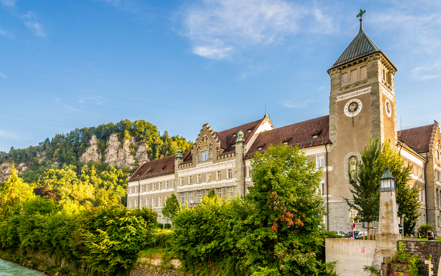 Historisches Gebäude an einem Fluss mit grünen Bäumen und Hügel im Hintergrund.