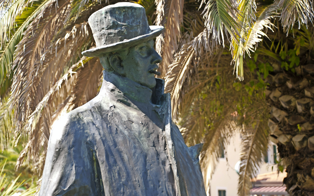 Statue eines Mannes mit Hut vor Palmen im Hintergrund.