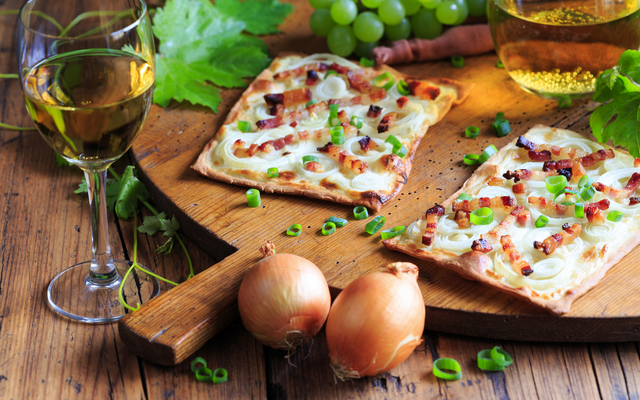 Flammkuchen mit Speck und Zwiebeln auf Holzbrett, daneben Weißwein und Trauben.