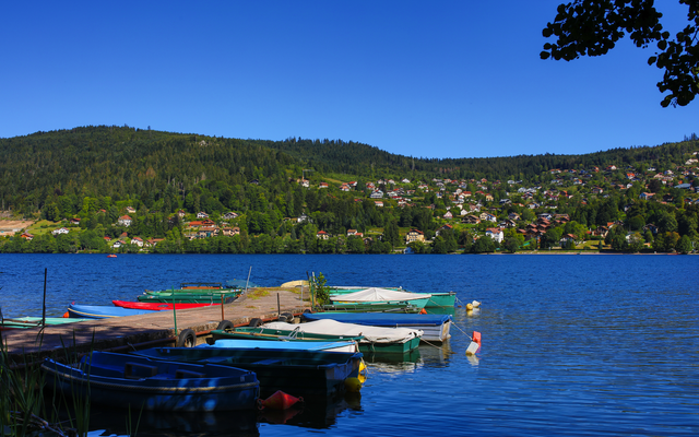 Boote am Steg vor einem See mit Häusern und Wäldern im Hintergrund