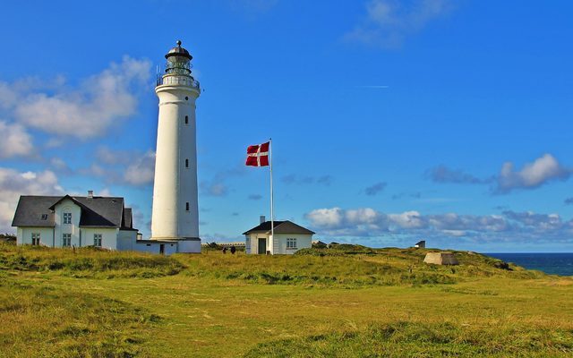 Leuchtturm mit dänischer Flagge vor blauem Himmel und Meer im Hintergrund.