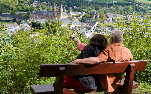 Älteres Paar sitzt auf Bank und blickt auf Stadt und Kirche in hügeliger Landschaft.