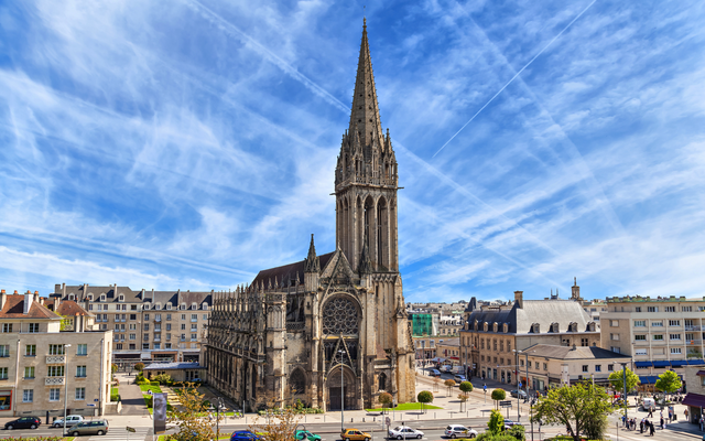 Gotische Kirche mit hohem Turm vor blauem Himmel in einer Stadtumgebung.