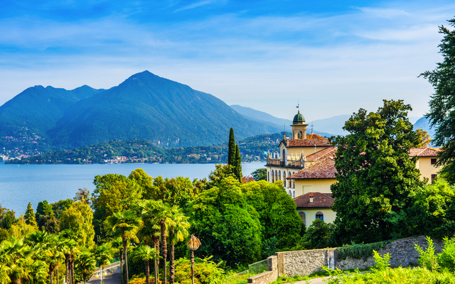 Landschaft mit Palmen, Villa und Blick auf einen See und Berge im Hintergrund.