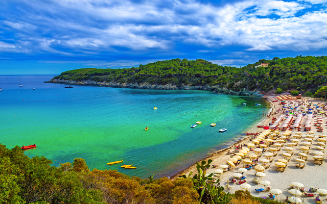 Strand mit Sonnenschirmen und türkisfarbenem Meer vor bewaldeten Hügeln unter blauem Himmel
