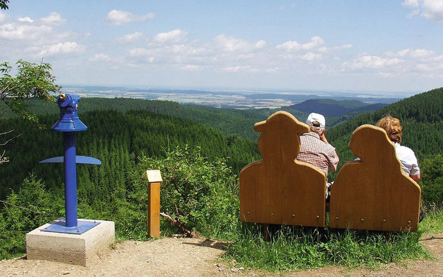Personen auf zwei Bänken mit Aussicht auf bewaldete Hügel und Fernglas.